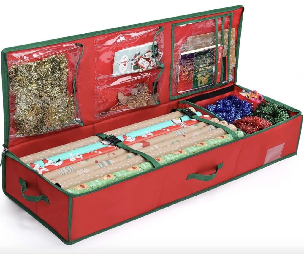 Open red storage container for holiday wrapping supplies with multiple compartments, holding rolls of wrapping paper, ribbon, tape, and various decorations like tinsel and bows. Perfect for organizing Christmas crafts for adults, with transparent pockets showing small gift tags and cards.